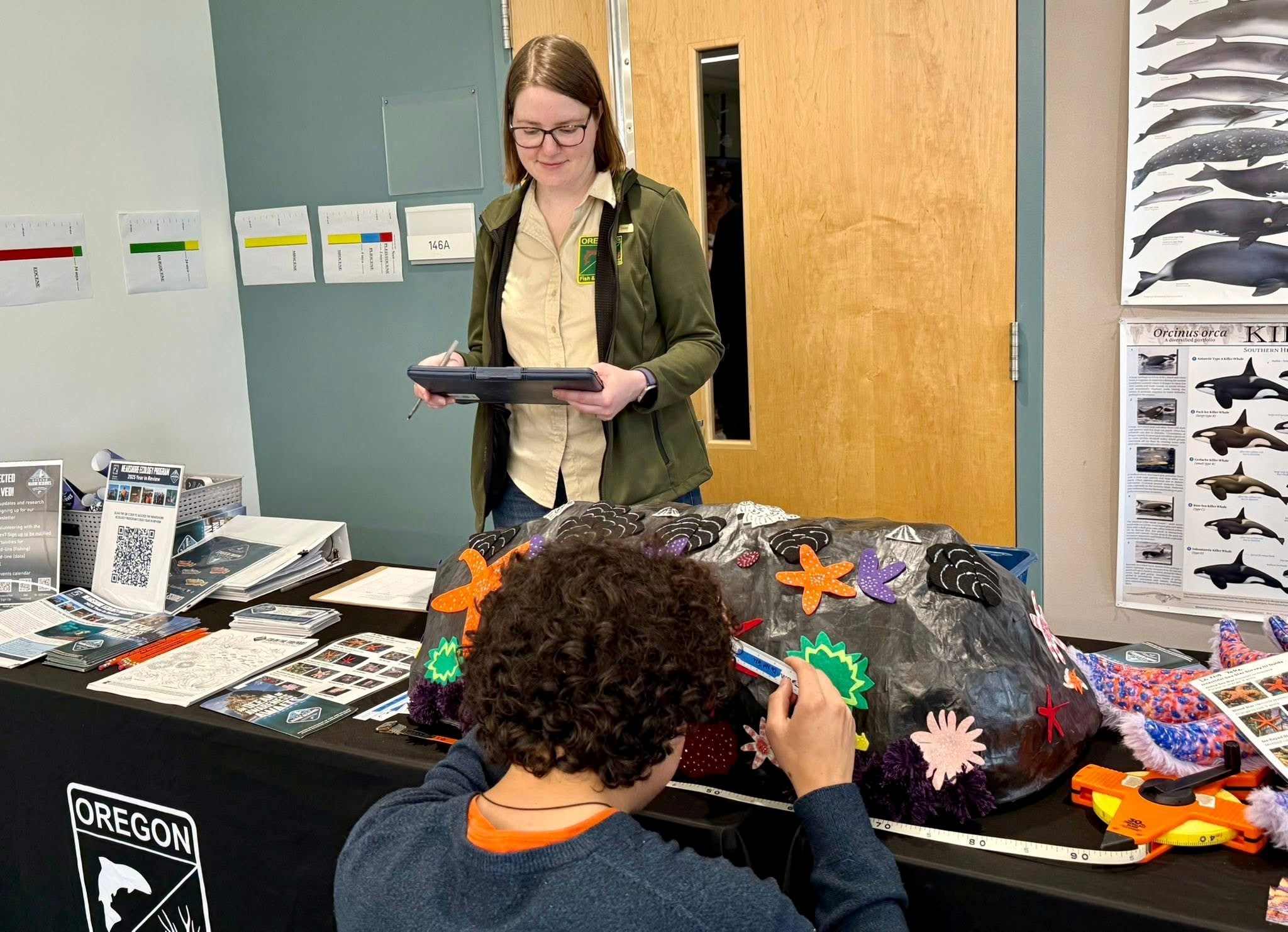Person measures arm of felt sea star on a paper mache rock while another person holding a clipboard records the data at Marine Science Day 2026.
